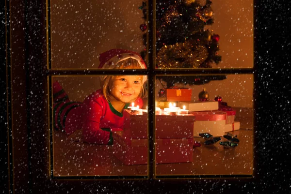 Happy cheerful little girl blowing candles at Christmas, view from outside window — Stock Photo, Image
