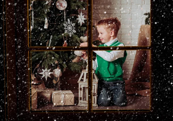 Little boy decorating Christmas tree viewed through window from outside — Stock Photo, Image