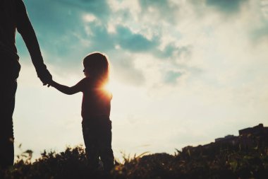 Gün batımında ebeveyn elini tutan küçük kızın silueti.