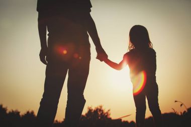 Gün batımında ebeveyn elini tutan küçük kızın silueti.