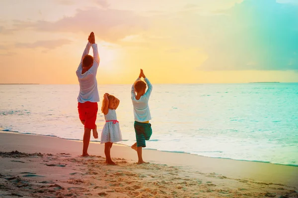 Отец с сыном и дочерью занимаются йогой на пляже на закате — стоковое фото