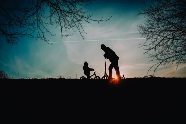 Silhouette de père et petite fille à vélo et scooter au coucher du soleil — Photo