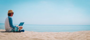 Uzaktan kumandalı iş - genç adam sahilde dizüstü bilgisayarla çalışıyor