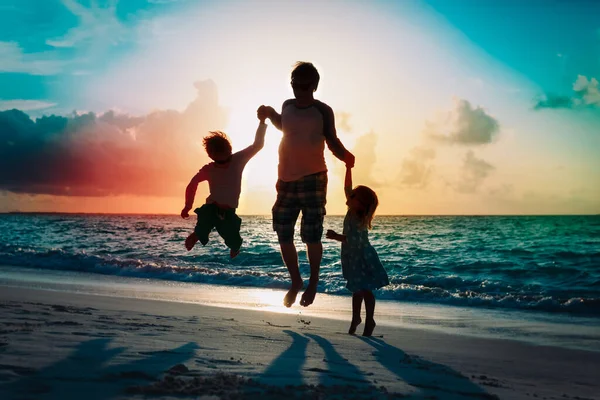 Gelukkig vader met kinderen springen van vreugde bij zonsondergang strand — Stockfoto