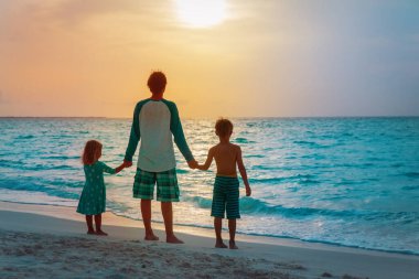 Baba ve çocuklar gün batımında sahilde yürüyorlar.