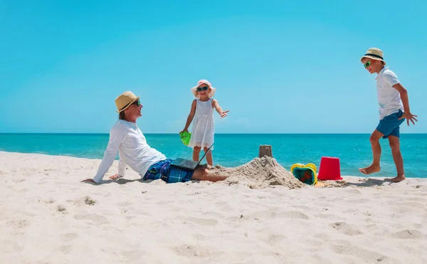 Vader met laptop proberen te werken en kinderen spelen met zand op het strand — Stockfoto