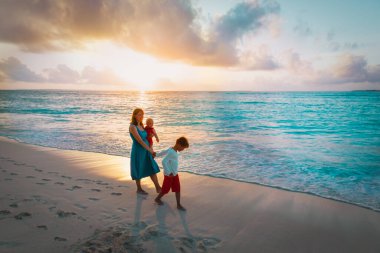 Anne ve çocuklar gün batımında sahilde yürüyorlar.