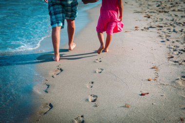 Baba ve kızı kumsalda yürüyor. Kumda ayak izleri bırakıyor.