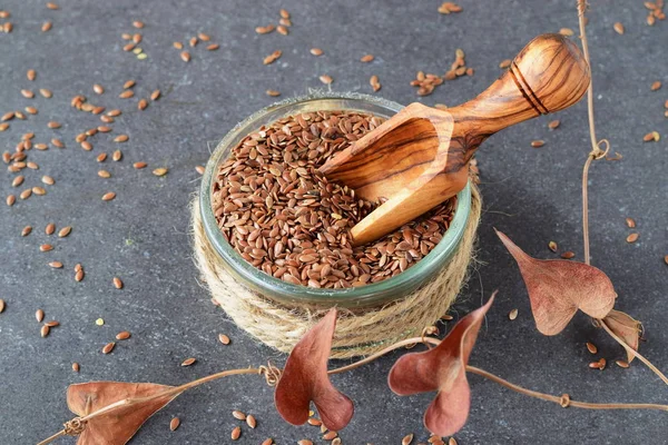 A glass bowl of flax seeds with olive scoop surrounded by artificial flowers on a grey abstract background. Healthy eating concept. — Stock Photo, Image