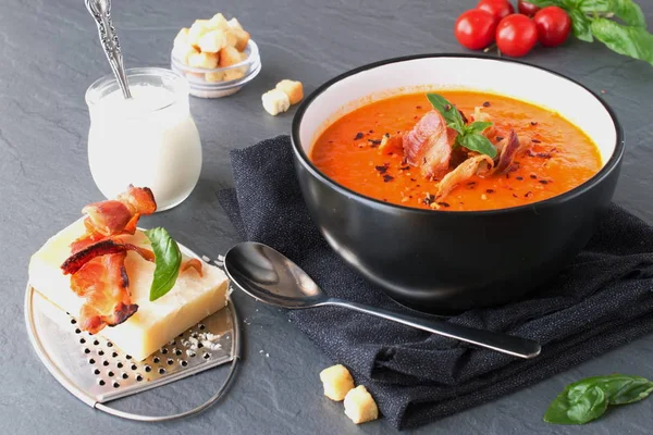 Толстый томатный суп с базиликом и жареным беконом в черной керамической миске на сером абстрактном фоне. Концепция здорового питания . — стоковое фото