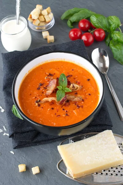Толстый томатный суп с базиликом и жареным беконом в черной керамической миске на сером абстрактном фоне. Концепция здорового питания . — стоковое фото