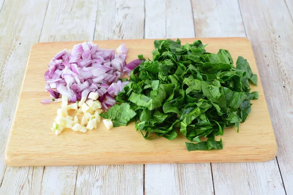 Step by step cooking. Cut red onion, spinach and garlic on a wooden cutting board on a light wooden background. — Stock Photo, Image
