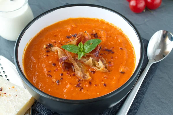 Толстый томатный суп с базиликом и жареным беконом в черной керамической миске на сером абстрактном фоне. Концепция здорового питания — стоковое фото