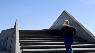 Çekici İskandinav, beyaz kadın, egzersiz yapmak için merdivenleri koşarak çıkıyor. Kamera ona yukarıdan bakıyor. Mavi gökyüzü ve bulutlarla güneşli güzel bir gün. Brian Holm Nielsen 'in stok video görüntüleri