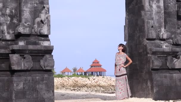 Jeune femme sexy dans une robe rester près des portes balinaises et admire la vue. Maison d'été asiatique et océan sur le fond. 1080p, 50 images par seconde, Full HD . — Video