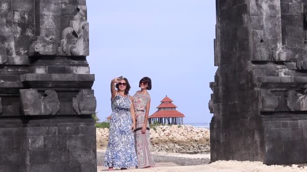 Две молодые довольно сексуальные дамы делают селфи рядом с балийским храмом, океаном и летним домом на заднем плане. Улыбаясь и смеясь. Захватывающее путешествие на Бали. Автопортрет, прекрасный вид . — стоковое видео
