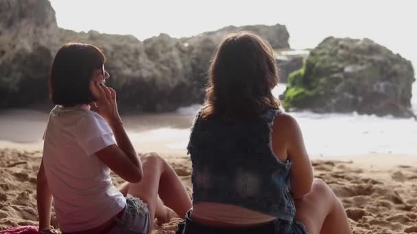 Красивые женщины в очках сидят на пляже возле океана, разговаривают, смеются и улыбаются. Одна женщина использует смартфон. Отдых девочек на острове Бали, Индонезия. Лучшие друзья. Вид сзади . — стоковое видео