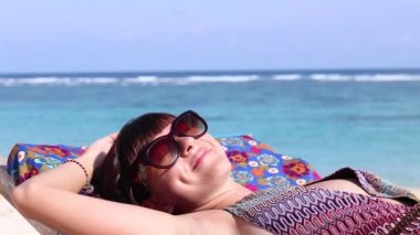 Yakın çekim genç mutlu kadın yatıyor ve güzel plajda şezlong üzerinde güneşleniyor, gülümsüyor ve hayal. Bali, Endonezya. Okyanus arka plan, sakin dalgalar ve güneş, rahatlayın. Full Hd, 50 fps, 1080p.