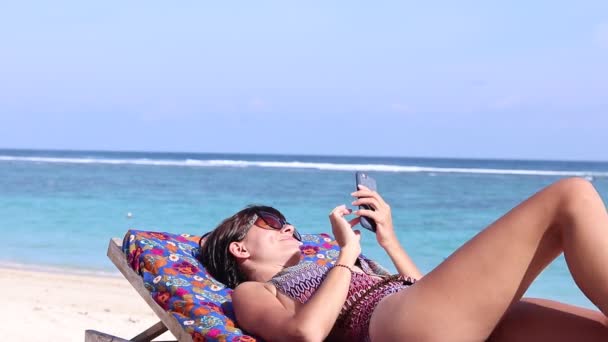 スマートフォンと笑顔を使用して美しい熱帯のビーチでセクシーな魅力的な若い白人女性。デッキチェアでリラックスした女性の日光浴。ビーチで女性の健康的なウェルネスライフスタイルにフィット. — ストック動画