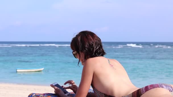 Sexy přitažlivá mladá Kavkazská žena na nádherné tropické pláži používající smartphone a usmívající se. Uvolněná Dámské lázně na palubě židle. Přizpůsobení zdravé wellness životní stylu žen na pláži. — Stock video