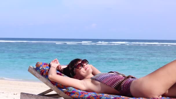 Close-up young happy woman laying and sunbaths on the deck chair on the beautiful beach, smiling and dreaming. Bali, Indonesia. Ocean background, calm waves and sun, relax. Full HD, 50 fps, 1080p. — Stock Video