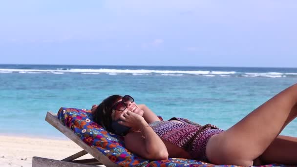 Sexy attractive young caucasian woman on the beautiful tropical beach using smartphone and smiling. Relaxed lady sunbaths on the deck chair. Fit female living healthy wellness lifestyle on the beach. — Stock Video