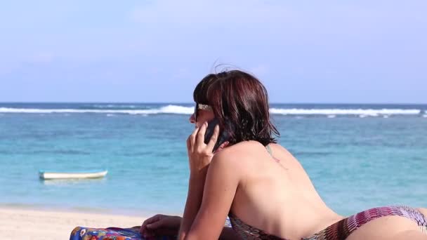 Sexy atraente jovem caucasiana na bela praia tropical usando smartphone e sorrindo. Banhos de sol relaxados na cadeira de praia. Apto feminino vivendo um estilo de vida saudável na praia . — Vídeo de Stock