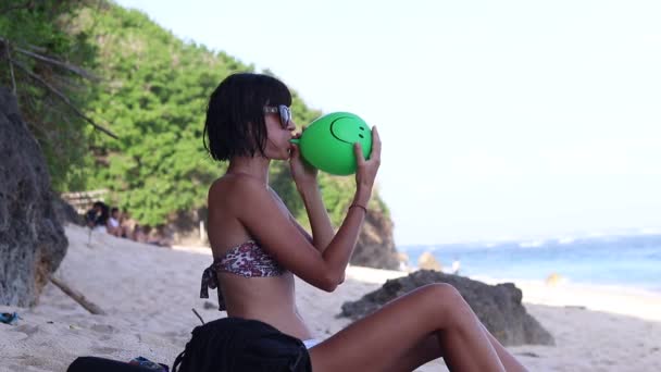 Σέξι νεαρή γυναίκα φυσώντας φουσκώνοντας ένα πράσινο συννεφάκι, νεαρή γυναίκα στην παραλία. Ωκεανός, Μπαλί, Ινδονησία. — Αρχείο Βίντεο