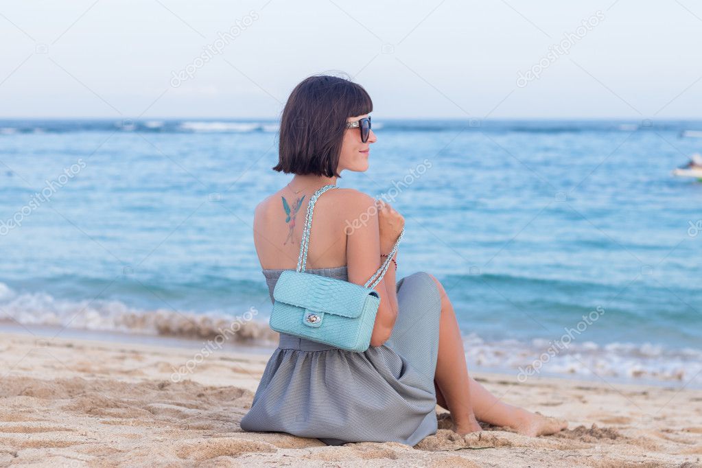 Fashionable young woman with tattoo on her back in black and white clothes with a luxury snakeskin handbag on the beautiful beach of tropicsl island Bali, Indonesia. Nusa Dua area. Amazing ocean view.