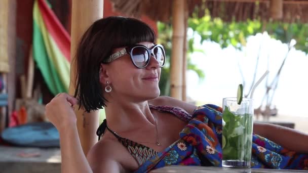 Красивая брюнетка в солнечных очках лежит в беседке с коктейлем в солнечный день. Остров Бали, Амед, Индонезия . — стоковое видео