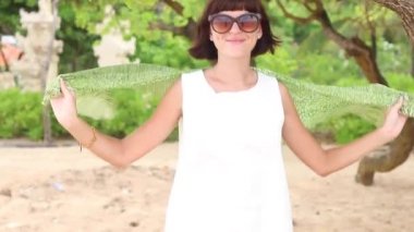 Yeşil İpek eşarp ve gözlük olan mutlu kadın tropikal Bali Adası, Endonezya kumsalda döne döne. Ağır çekim. Ağaçlar ve avuç içi arka planda.