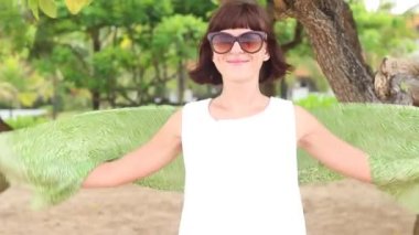 Yeşil İpek eşarp ve gözlük olan mutlu kadın tropikal Bali Adası, Endonezya kumsalda döne döne. Ağır çekim. Ağaçlar ve avuç içi arka planda.