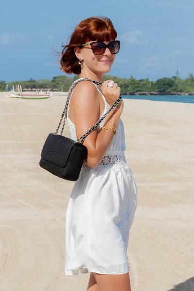 Sexy Mode brünettes Model in weißem Kleid posiert am Strand. mit Sonnenbrille und Luxus-Handtasche aus Schlangenhaut-Python. bali, indonesien. — Stockfoto