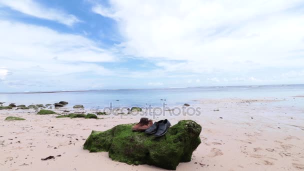 Scarpe vecchie sulla grande pietra con muschio. Piccole rocce sparse sulla sabbia della spiaggia da vicino. Bellissimo paesaggio oceanico, cielo incredibile. Bali, Indonesia . — Video Stock