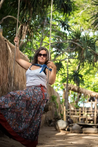 Sensual chica tierna sentada en un columpio con hermoso vestido. Tiene el pelo largo y oscuro. El columpio está en la playa con árboles verdes. Isla tropical de Bali, Indonesia . — Foto de Stock