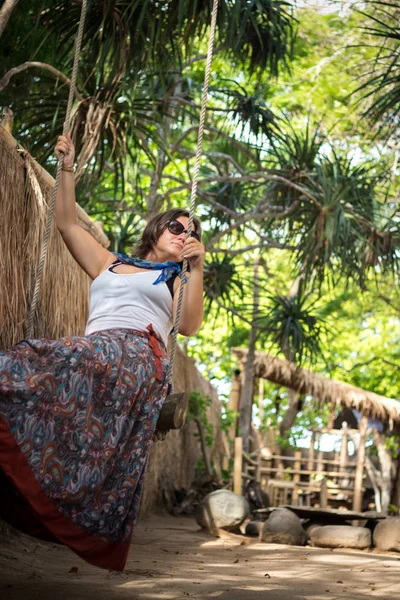 Sensual chica tierna sentada en un columpio con hermoso vestido. Tiene el pelo largo y oscuro. El columpio está en la playa con árboles verdes. Isla tropical de Bali, Indonesia . — Foto de Stock