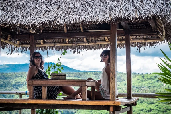 Dvě krásné šťastné mladé dívky sedí v dřevěný altán na slunečný den. a baví, úsměv a smích. Tropický ostrov Bali, Indonésie. — Stock fotografie