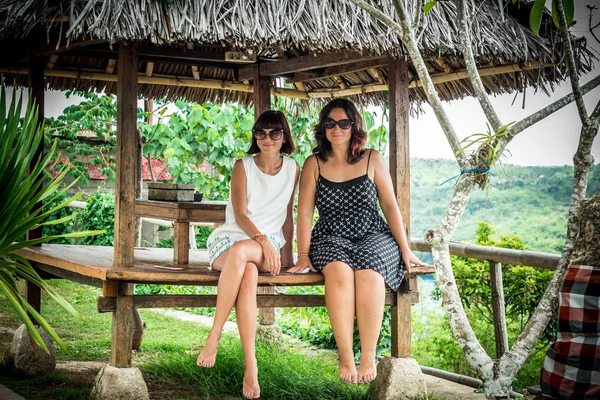 Dvě krásné šťastné mladé dívky sedí v dřevěný altán na slunečný den. a baví, úsměv a smích. Tropický ostrov Bali, Indonésie. — Stock fotografie