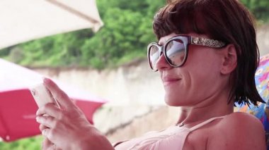 Kumsalda gülümseyen mobil akıllı telefon kullanan kadın. Güzel Asyalı beyaz kadın modeli bir tropik adada Bali, Endonezya.