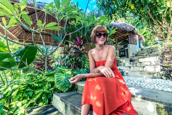 Jonge modieuze vrouw in rode jurk in een tropische tuin. Portret van gelukkige vrouw ontspannen op Bali eiland, Indonesië. — Stockfoto