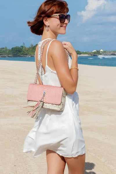 Сексуальная модель брюнетки в белом платье позирует на пляже. Ношение солнцезащитных очков и роскошной сумочки из змеиной кожи питона ручной работы. Бали, Индонезия . — стоковое фото