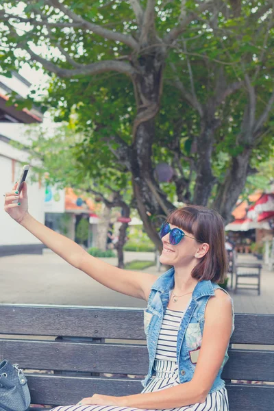 Parkta bir bankta oturmuş smartphone kullanan güzel genç seksi kadın. Bali Adası, Endonezya.