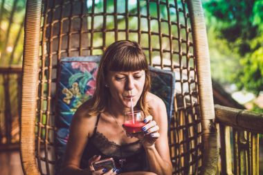 Güzel kadın balkonunda yağmur ormanları villada salıncakta otururken karpuz jucie içme. Bali, Endonezya'nın tropik ada.