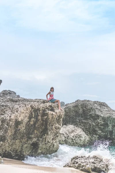 海岸の石の上に座って美しい若い女性。インドネシア、バリ島の熱帯の島. — ストック写真