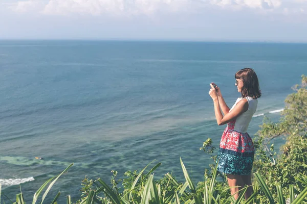 Młoda kobieta, robienia zdjęć na klifie, z tle piękny ocean w słoneczny dzień. Wyspa Bali. — Zdjęcie stockowe