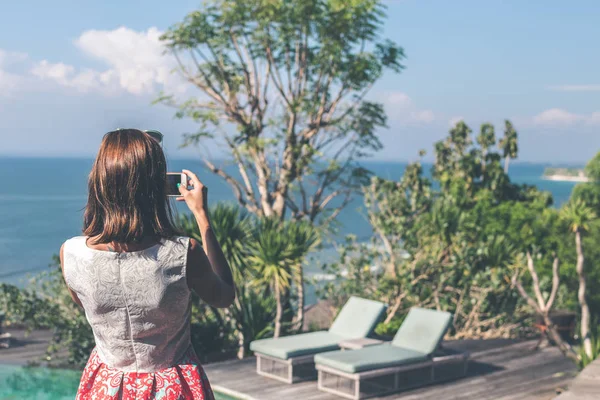 Jonge vrouw nemen van foto's op de klif met een prachtige oceaan achtergrond op zonnige dag. Bali eiland. — Stockfoto