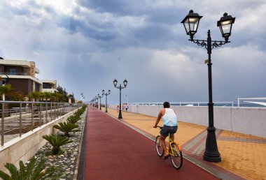 Sochi (Adler), Rusya Federasyonu - 22 Ağustos 2017: Turist binicilik bisiklet kauçuk kaplamalı Bisiklet şeritli fenerler ve genç palmiye ağaçları önünde Arfa Park-Hotel Olympic Park yakınındaki waterfront alan gezinti yolu üzerinde