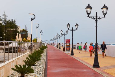 Sochi (Adler), Rusya Federasyonu - 22 Ağustos 2017: fenerler ve genç palmiye ağaçları önünde Arfa Park-Hotel Sochi Olympic Park yakınındaki güzel sahil yürüyen turist gezi
