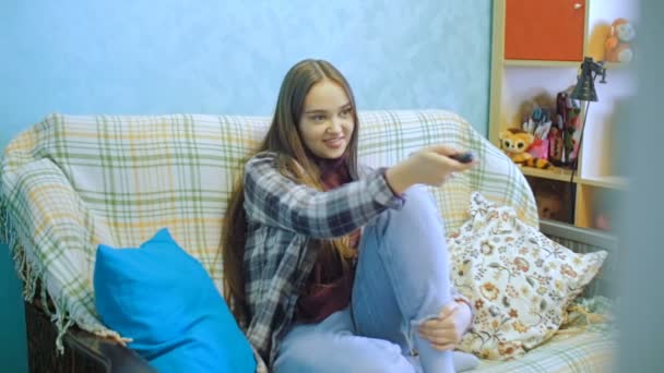 La joven está sentada en el sofá y viendo la tele. La mujer cambia los canales de televisión . — Vídeos de Stock