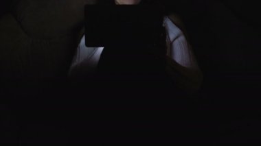 Beyaz bir ceketi giyen bir kadın onun elinde bir tablet ile karanlıkta oturmuş.